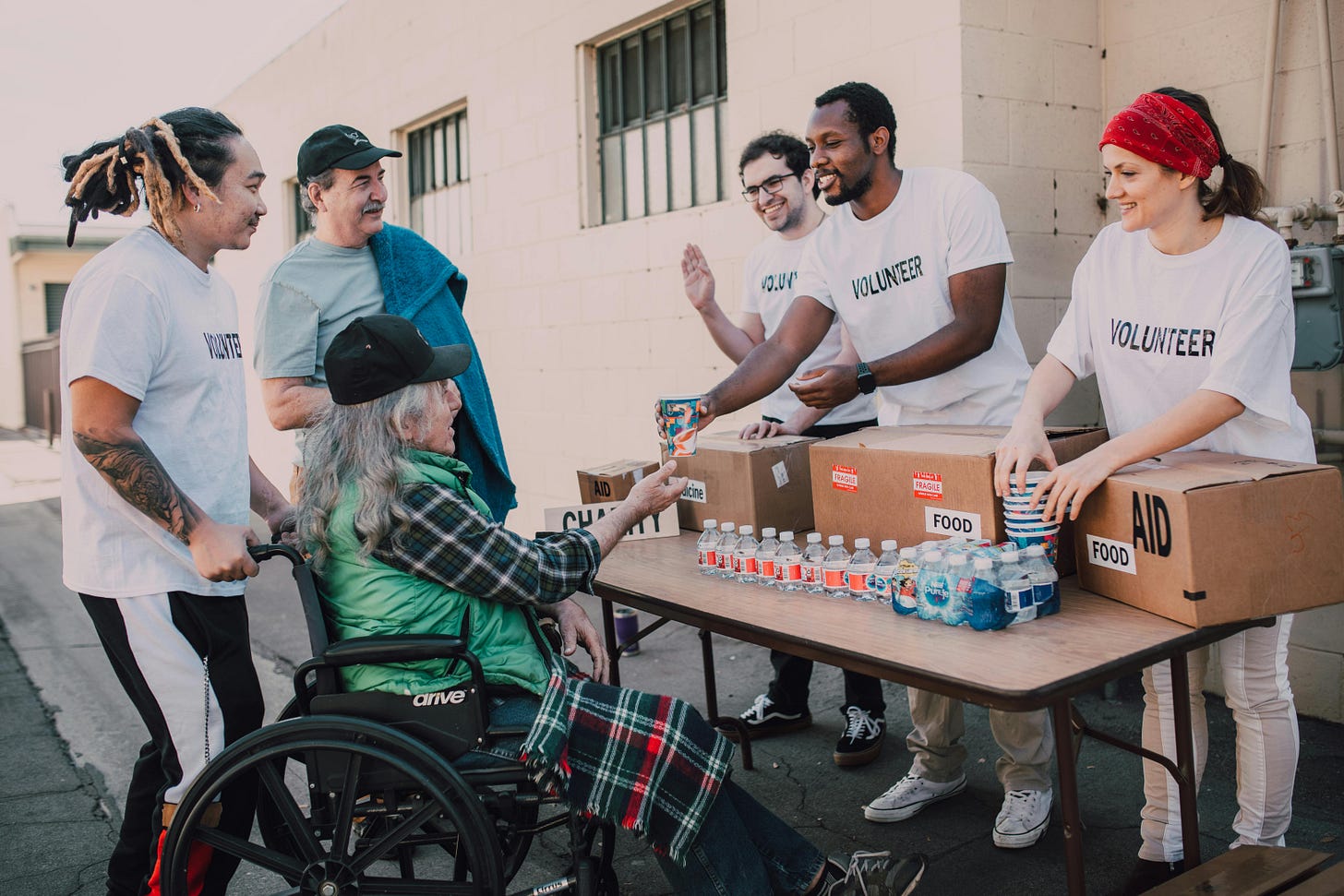 Three people standing at a table wearing white t-shirts with the word "volunteer" in black. They are talking to three people on the other side of the table. One is in a wheelchair wearing a green vest and plaid shirt, another is wearing a white shirt that says "Volunteer" and another is in a blue shirt and black hat. The table has boxes of food and bottled water on it.