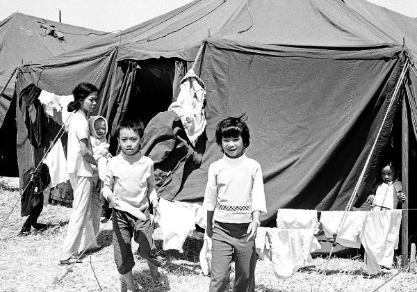 Vietnamese refugee families, including women and children, walk past rows of tents in a temporary resettlement camp in the United States after the fall of Saigon, 1975.