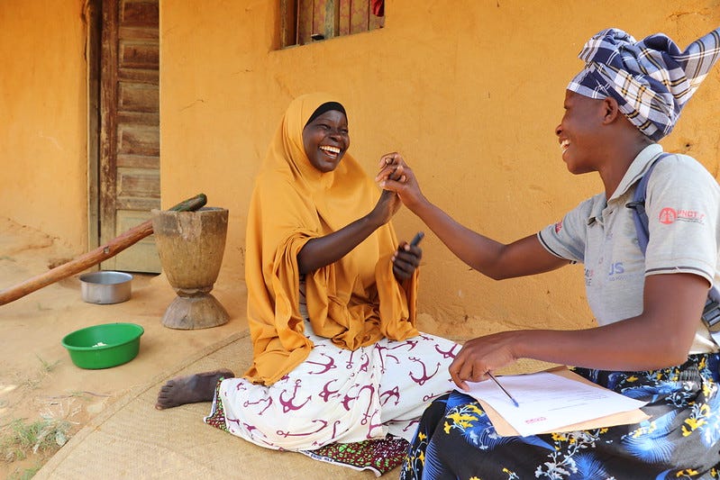 On a woven mat in front of a simple home with bright orange stucco walls, the homeowner, a woman in a bright orange hijab (the shade almost matches the wall behind her!) and white skirt clasps hands with a woman wearing a grey polo shirt with the logo of a local aid organization, a bright blue and black skirt, and a plaid head scarf. In her lap are some papers; she holds a ballpoint pen in her free hand. Both women grin widely; their happiness is obvious. To the left of the pair can be seen an aluminum cook pot, a green plastic bowl, and a wooden mortar and long wooden pestle. The wooden front door of the house is slightly ajar, and just above the homeowner's head is a window with no glass, but fabric curtains. Balanced on the windowframe is a cell phone. The scene is in daylight, bright but without glare. On a woven mat in front of a simple home with bright orange stucco walls, the homeowner, a woman in a bright orange hijab (the shade almost matches the wall behind her!) and white skirt clasps hands with a woman wearing a grey polo shirt with the logo of a local aid organization, a bright blue and black skirt, and a plaid head scarf. In her lap are some papers; she holds a ballpoint pen in her free hand. Both women grin widely; their happiness is obvious. To the left of the pair can be seen an aluminum cook pot, a green plastic bowl, and a wooden mortar and long wooden pestle. The wooden front door of the house is slightly ajar, and just above the homeowner's head is a window with no glass, but fabric curtains. Balanced on the windowframe is a cell phone. The scene is in daylight, bright but without glare.