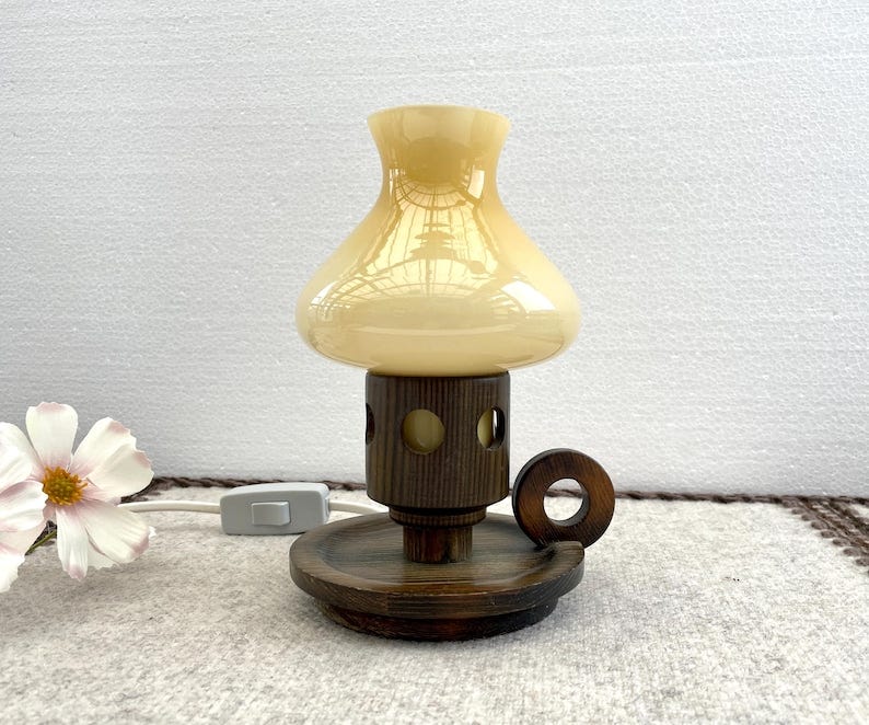 May include: A vintage table lamp with a mushroom-shaped, cream-colored glass shade. The lamp features a dark brown wooden base with a circular handle and a cylindrical body with circular cutouts. The lamp is plugged into a wall outlet. May include: A vintage table lamp with a mushroom-shaped, cream-colored glass shade. The lamp features a dark brown wooden base with a circular handle and a cylindrical body with circular cutouts. The lamp is plugged into a wall outlet.