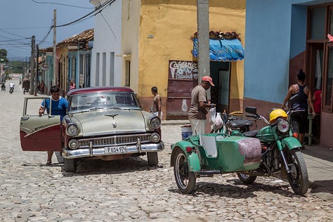 A 3x3 grid of nine photographs depicting life in Cuba. Images include: a boy diving into water; children sitting by a wall; flamingos in a lake; colorful colonial street scenes with classic cars and horse-drawn carts; a cat and a dog in a window; and a lamp-lit cobblestone street at night.