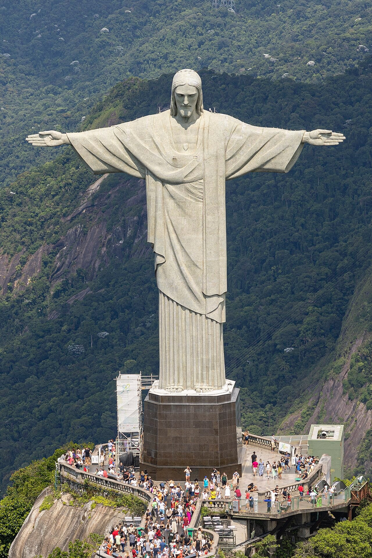 The white statue of Christ the Redeemer with arms spread wide on the summit of Corcovado, visitors crowding the platform at its base and green slopes below The white statue of Christ the Redeemer with arms spread wide on the summit of Corcovado, visitors crowding the platform at its base and green slopes below