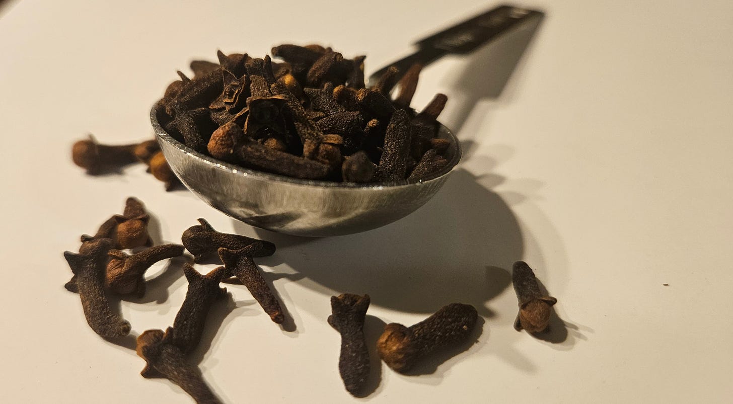 A heap of whole, dried organic clove buds spilling onto a clean white surface from a tablespoon