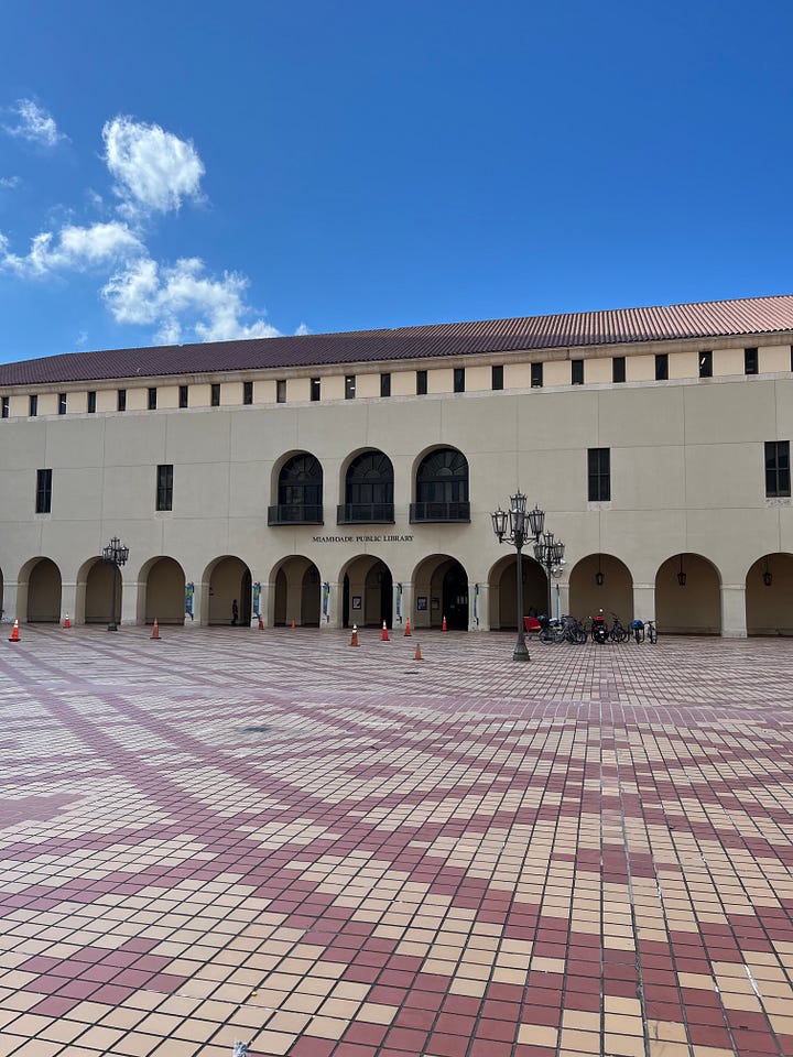 top left: exterior of Miami Pubic Library with Spanish tile courtyard; top right: view from second floor mezzanine looking down at the lobby of the Miami Public Library with orange tile floors and a central information desk; bottom left: books on the library shelf by Micol Ostow; bottom right: some art and special collections on the second floor