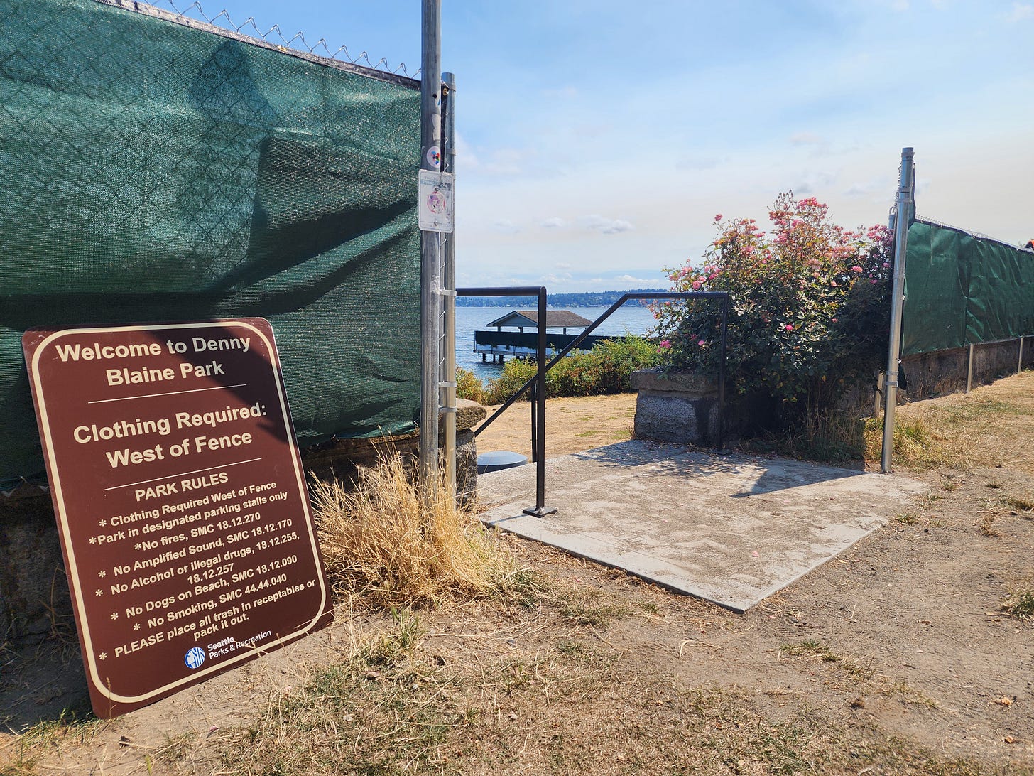A chain-link fence with green privacy tarp divides Denny Blaine Park, with a sign reading “Clothing Required: West of Fence” beside a narrow concrete stairway leading down toward Lake Washington and a small dock in the distance. A chain-link fence with green privacy tarp divides Denny Blaine Park, with a sign reading “Clothing Required: West of Fence” beside a narrow concrete stairway leading down toward Lake Washington and a small dock in the distance.