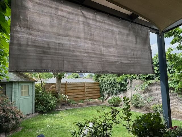A semi-transparent outdoor shade hangs from a pergola, partially obscuring a view of a well-kept garden with green grass, shrubs, a wooden shed, and a wooden privacy fence in the background.
