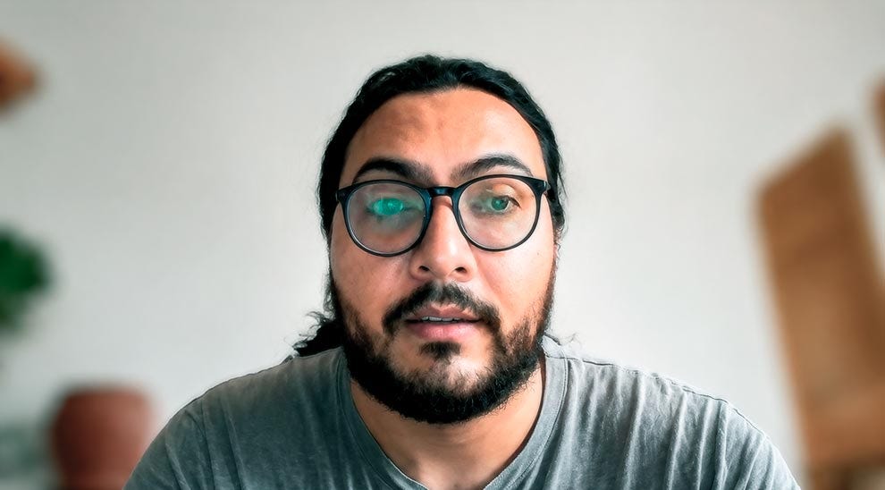 Pessoa usando óculos de armação preta e camiseta cinza, olhando diretamente para a câmera em ambiente interno com fundo claro e desfocado.