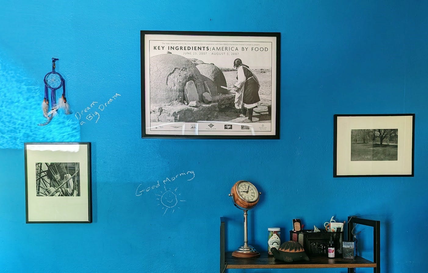 sky-blue chalkboard wall hung with art: a poster of woman baking in earthen oven at Acoma pueblo, flanked by photographs of a willow tree and a young woman posed reflectively behind the spokes of a wheel; above the latter is a small blue dream catcher; below the art is a bookshelf with clock and nicknacks