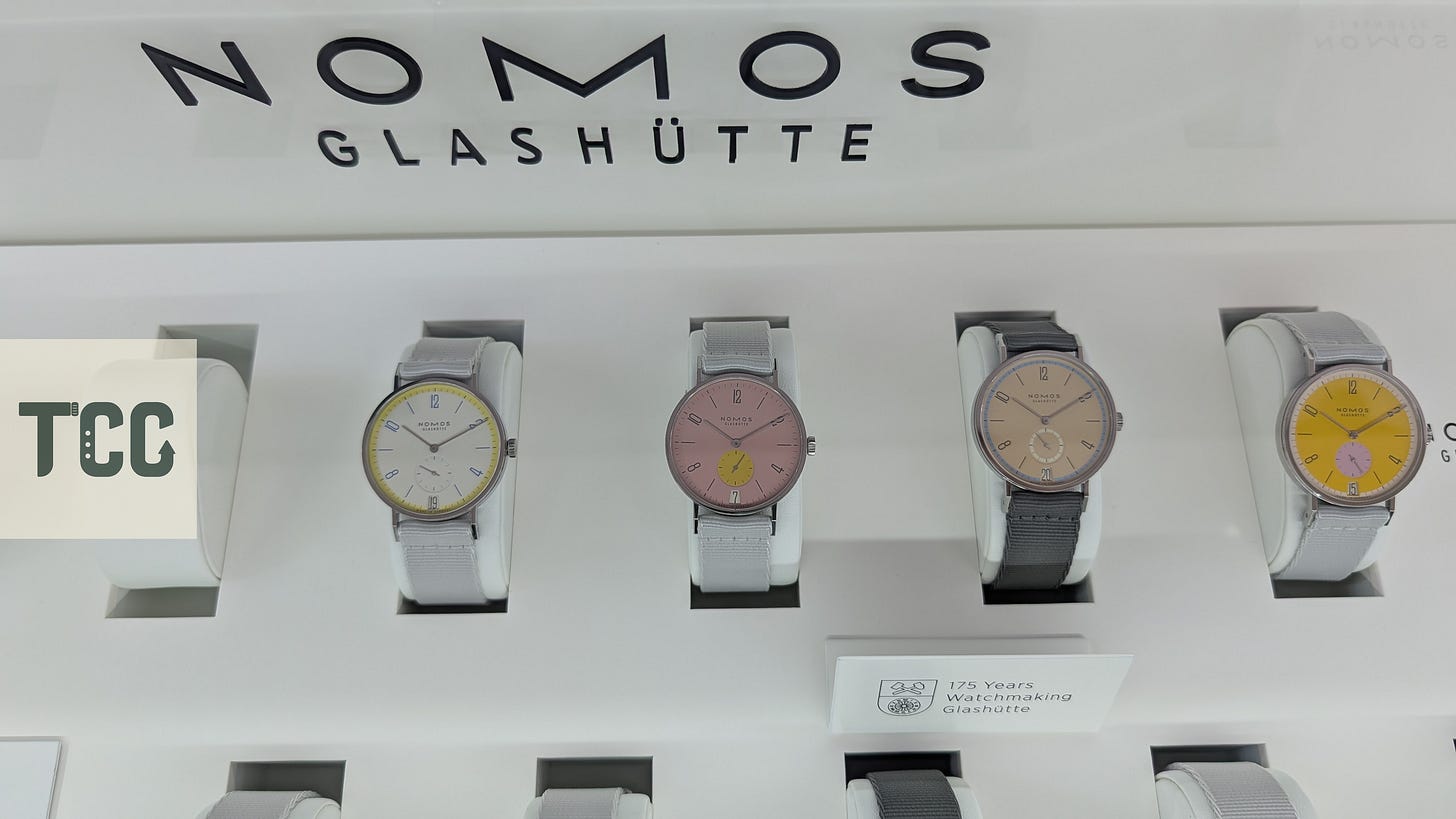 A close-up of four NOMOS watches in pastel and bold colorways—yellow, pink, tan, and mustard—each on display with light fabric straps beneath a sign reading “175 Years Watchmaking Glashütte.”