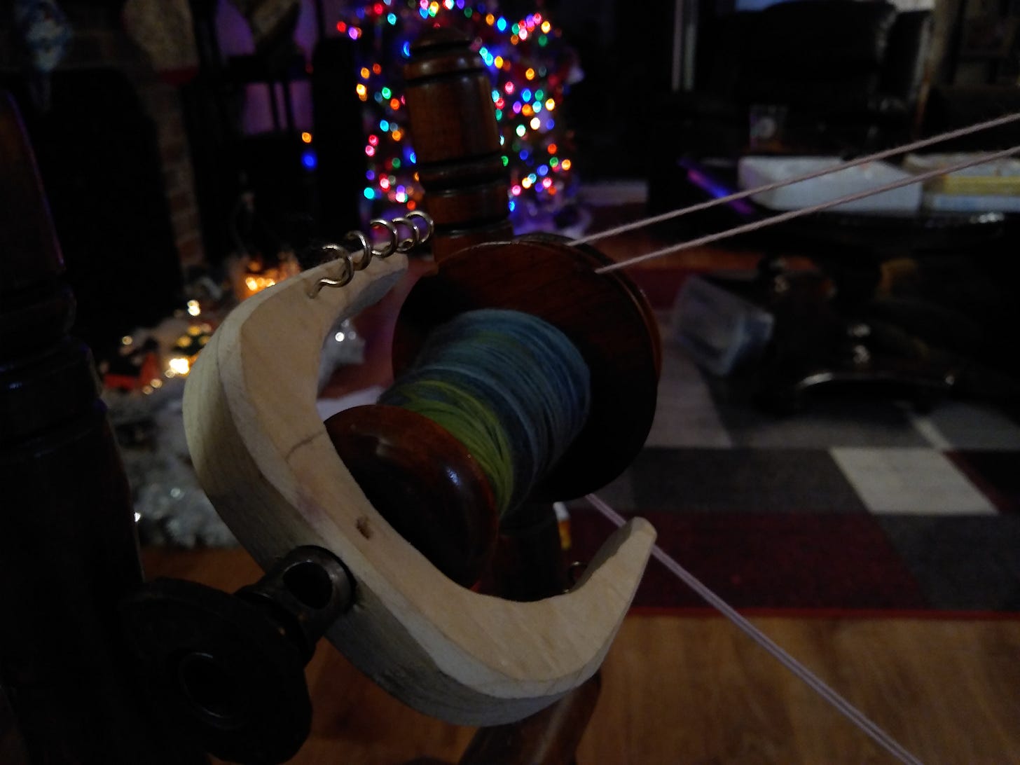 Dark picture of blue and green yarn on a bobbin on a spinning wheel, Christmas lights in the background