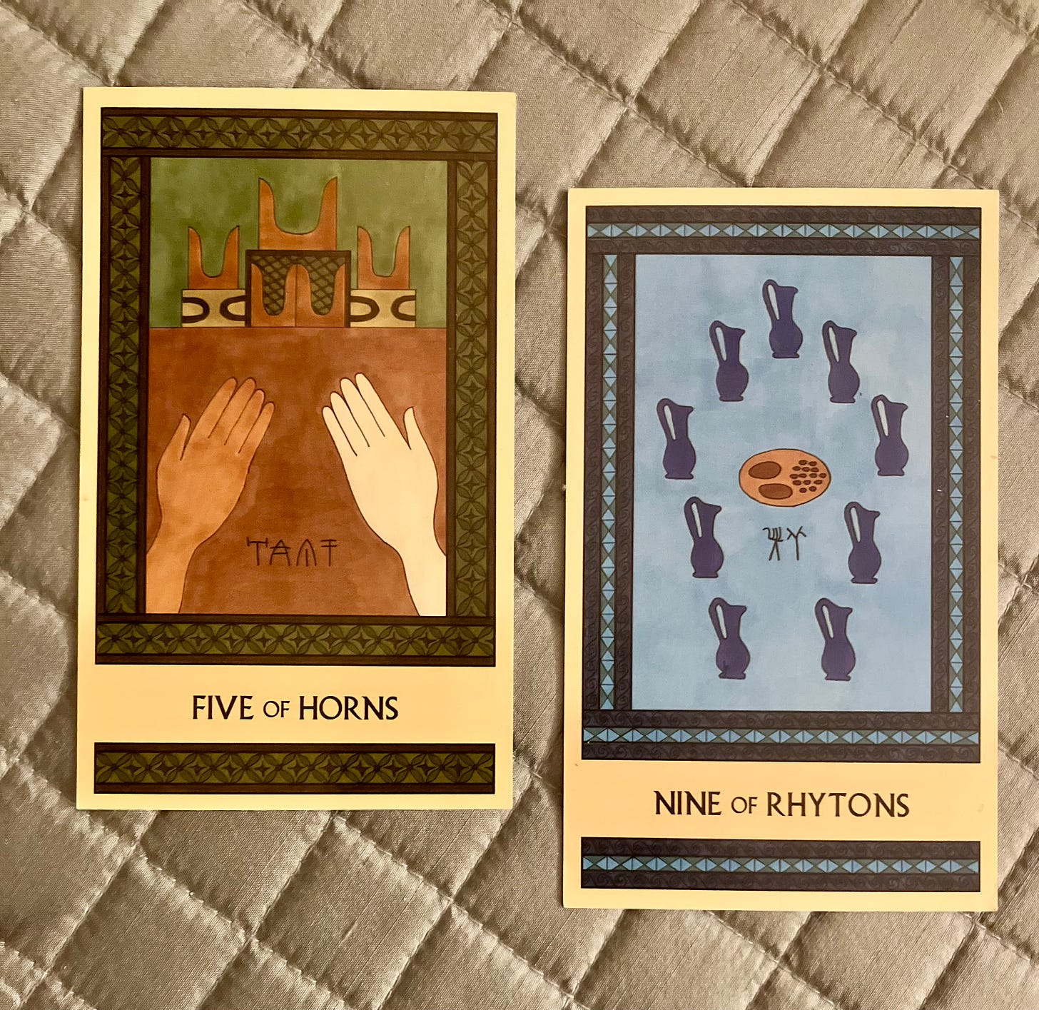 Two Minoan Tarot cards on a quilted silvery background. The Five of Horns is painted in shades of green and brown. It shows two hands reaching out toward a well-appointed shrine. The Nine of Rhytons is painted in shades of blue. It shows a platter of food surrounded by nine blue pitchers.