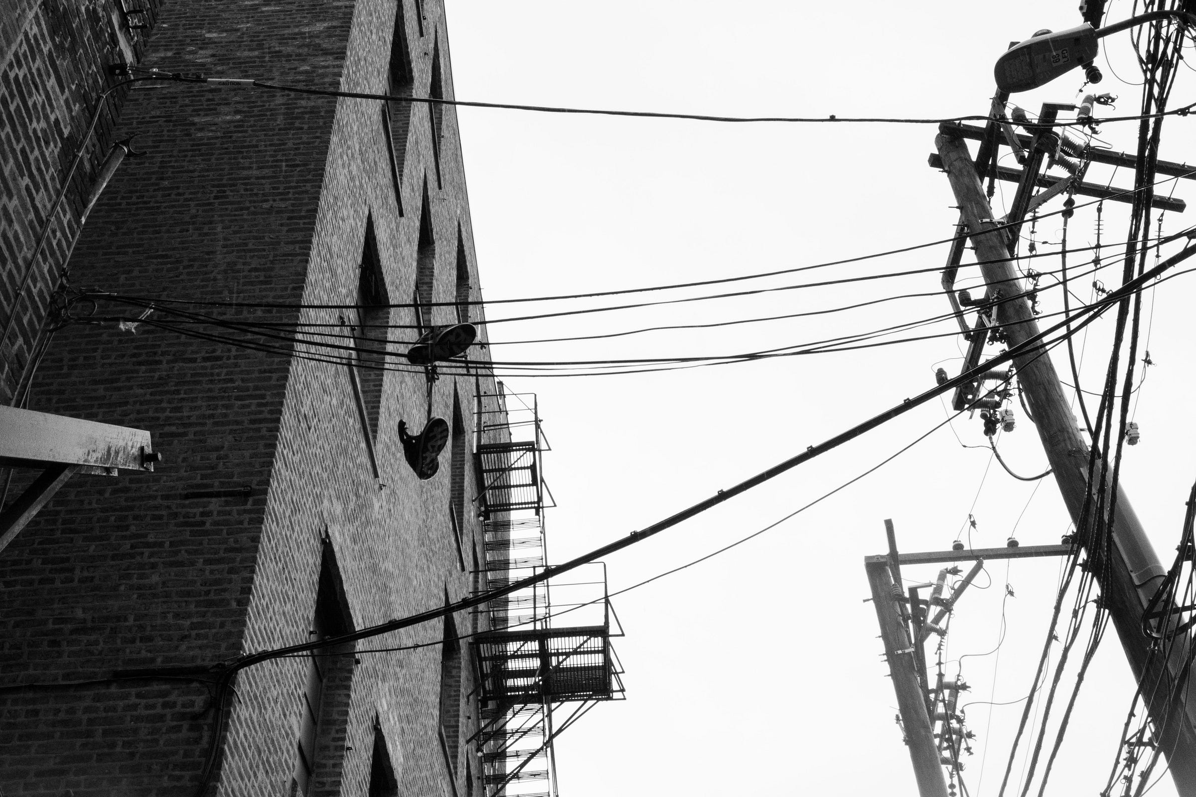 A pair of sneakers dangle from overhead power lines near a brick building with fire escapes, surrounded by a tangle of electrical wires and leaning utility poles. A pair of sneakers dangle from overhead power lines near a brick building with fire escapes, surrounded by a tangle of electrical wires and leaning utility poles.