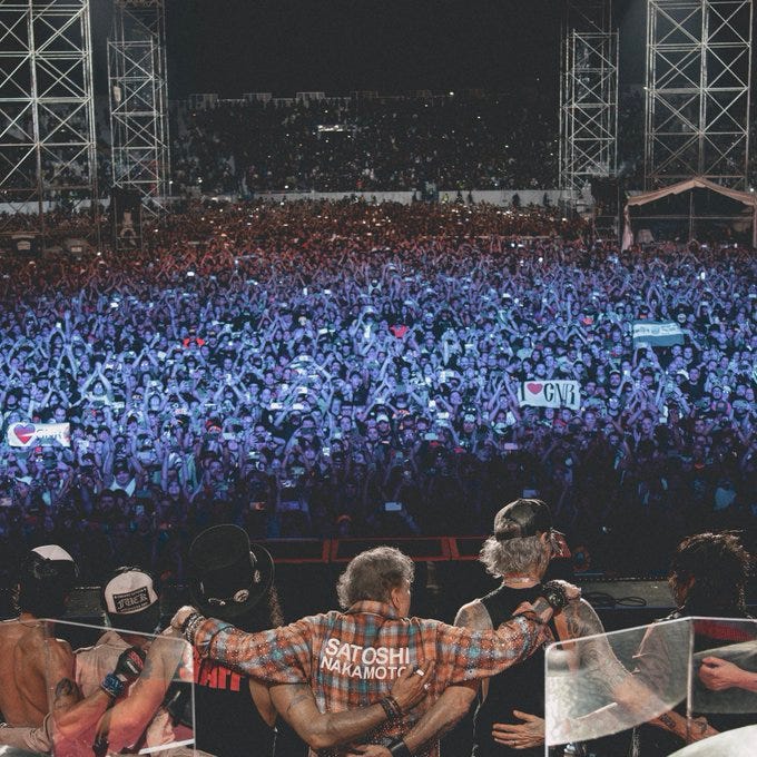 Large outdoor concert stage at night with metal framework and lighting rigs on both sides. Vast crowd of attendees waving arms and holding signs including one with a heart symbol. In foreground, several men on stage wearing casual clothing one with a shirt displaying Satoshi Nakamoto text others with band shirts and accessories like wristbands. Blue and purple lighting illuminates the scene with people in the background.