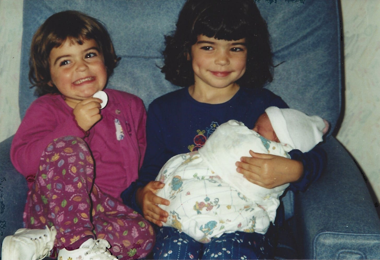 A 3-year-old girl sits next to her 5-year-old sister, who's cradling their newborn baby sister