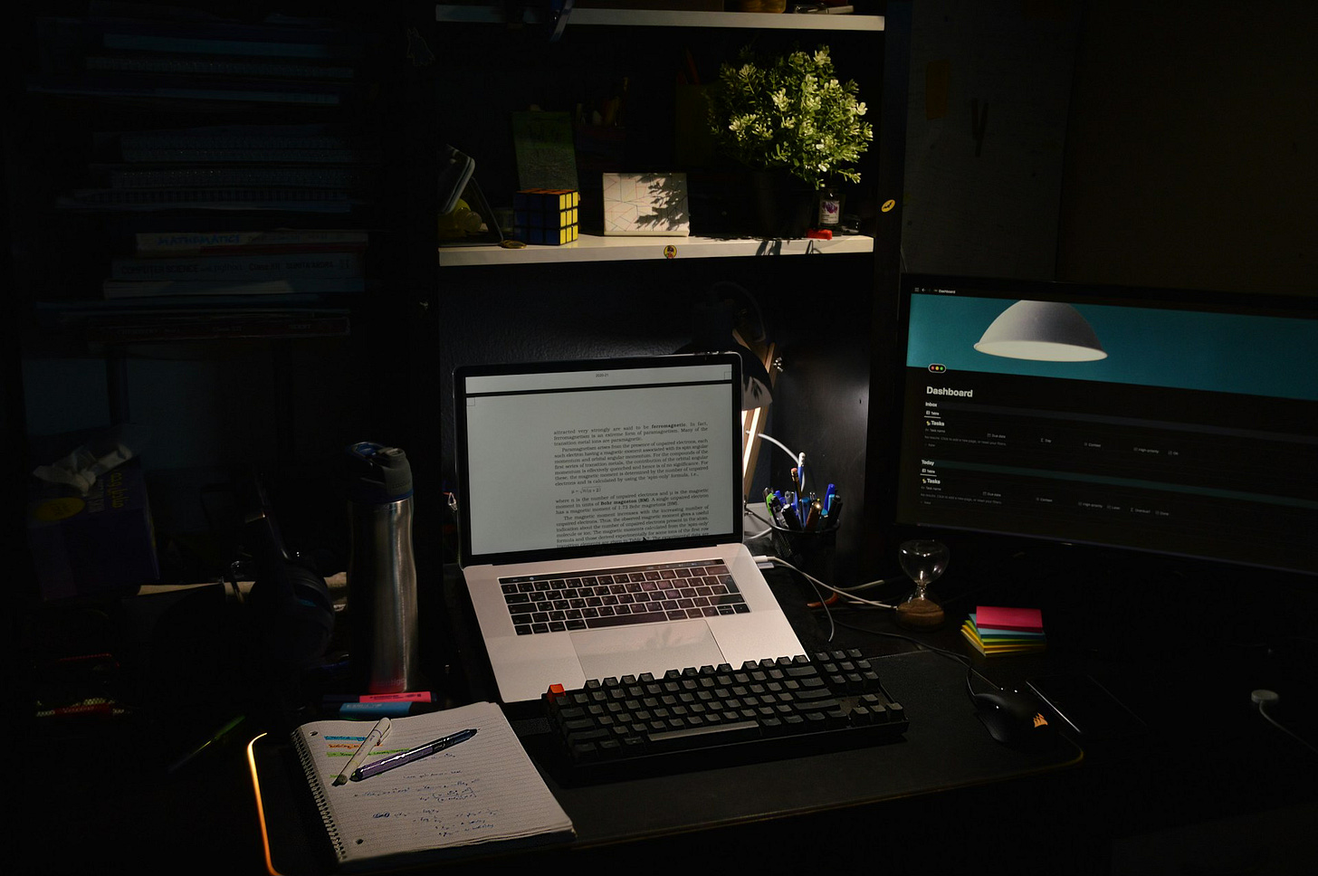 A workspace in a dark room. A black desk holds an open laptop propped on a stand, text filling the screen in a document. Beneath it rests a black keyboard with matching wrist rest, an open spiral notebook with notes and a few pens and highlighters, and a mouse. To one side is a metal water bottle, to the other a freestanding monitor open to a Notion dashboard. Above are several white shelves holding assorted items: Rubix cube, a plant, etc.