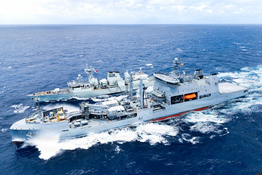 An aerial photograph shows two naval vessels conducting an underway replenishment (UNREP) operation at sea. The larger vessel, marked 'A11', appears to be a replenishment/auxiliary ship transferring supplies to a smaller warship alongside it. Both ships are underway in open ocean conditions with moderate sea state. An aerial photograph shows two naval vessels conducting an underway replenishment (UNREP) operation at sea. The larger vessel, marked 'A11', appears to be a replenishment/auxiliary ship transferring supplies to a smaller warship alongside it. Both ships are underway in open ocean conditions with moderate sea state.