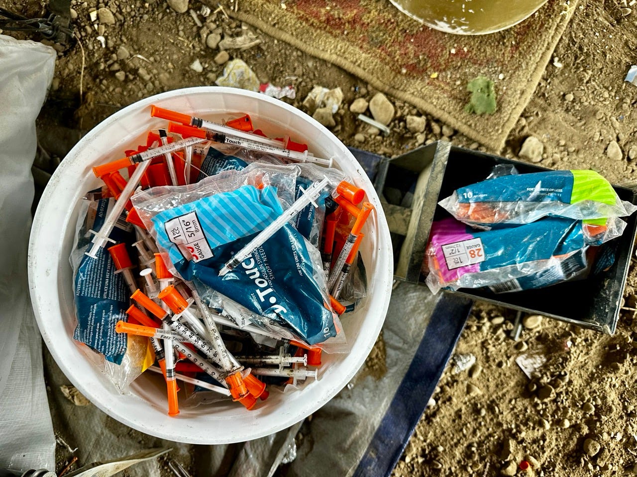 A bucket of syringes and other objects on the ground

Description automatically generated