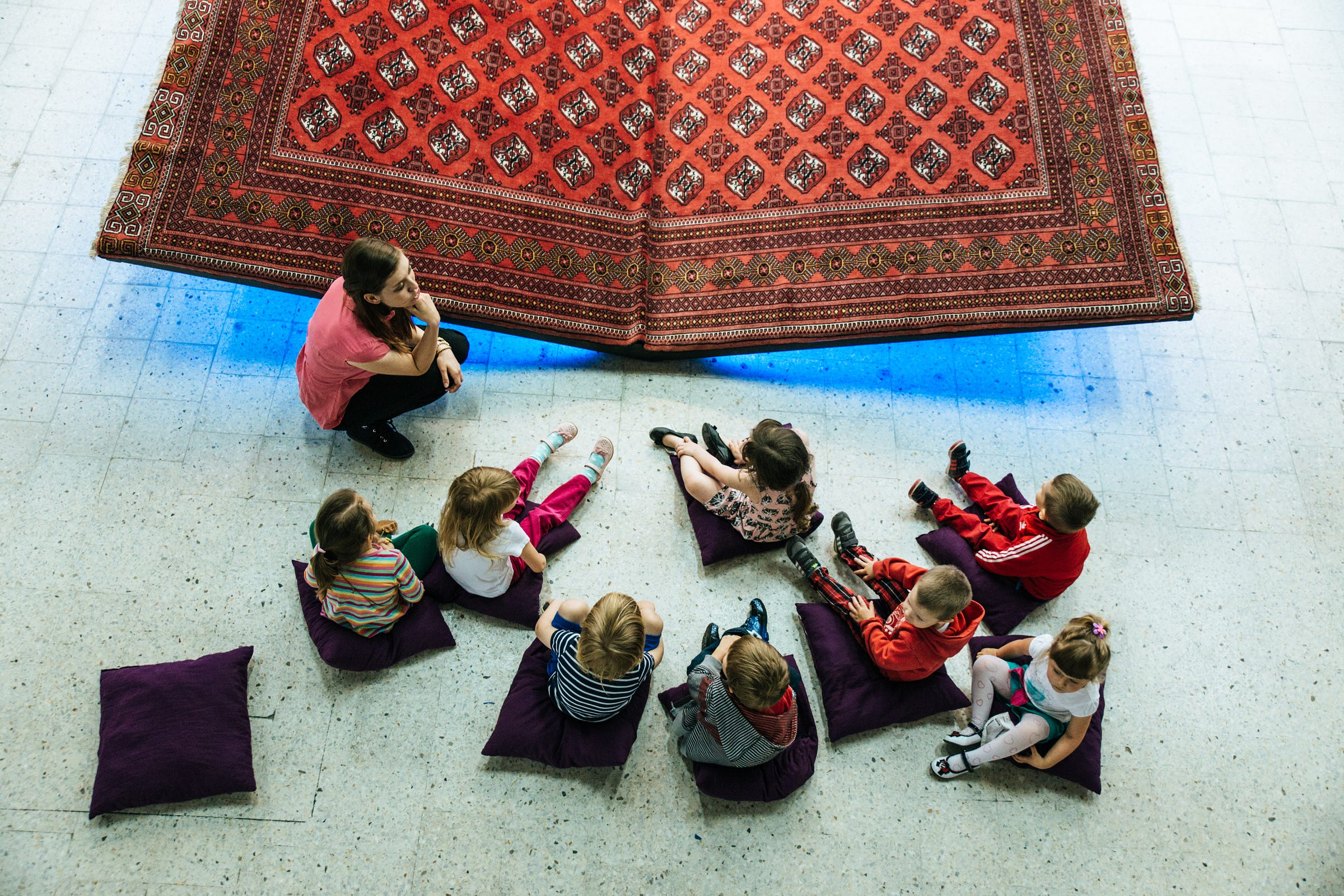 A young woman seated in front of a Persian carpet exhibit, as several young children look A young woman seated in front of a Persian carpet exhibit, as several young children look