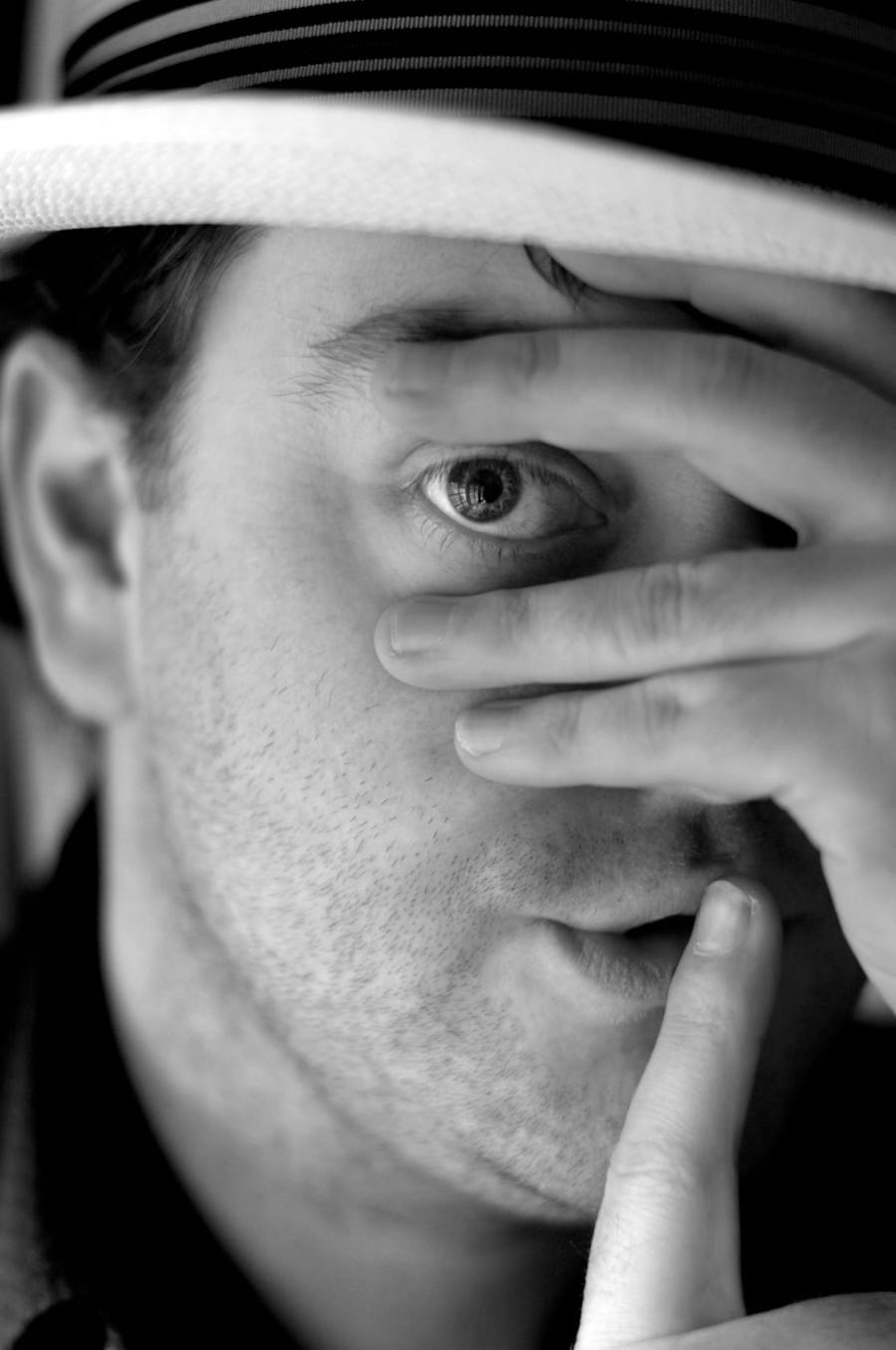 A portrait of someone in a straw hat masking their face with a hand and holding a finger up to their lips saying shush.