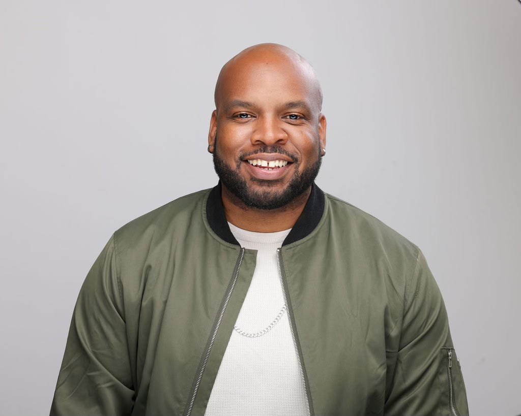 a headshot of Pastor Trey, a Black man with a bead who is smiling and wearing a white shirt with an olive green jacket