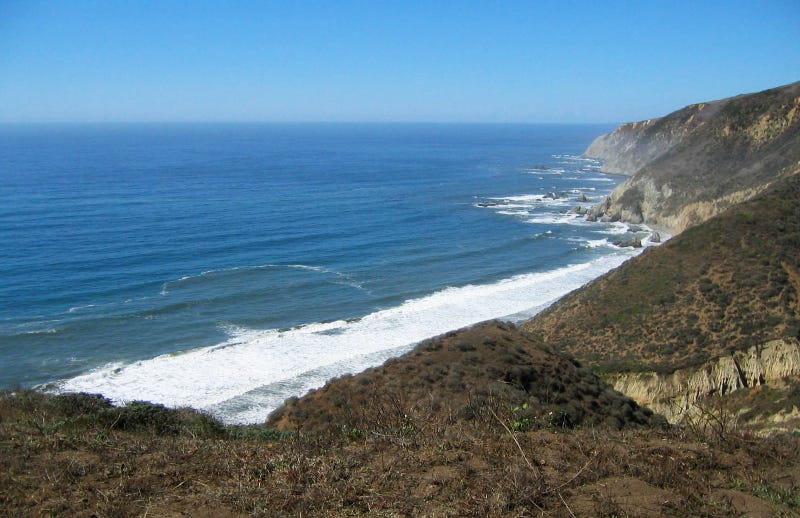 From a trail at the top of rocky seaside cliffs looking down toward the white foamy surf at the edge of the water. The deep blue rippling sea extends to forever, where it meets the lighter blue of the sky. From a trail at the top of rocky seaside cliffs looking down toward the white foamy surf at the edge of the water. The deep blue rippling sea extends to forever, where it meets the lighter blue of the sky.