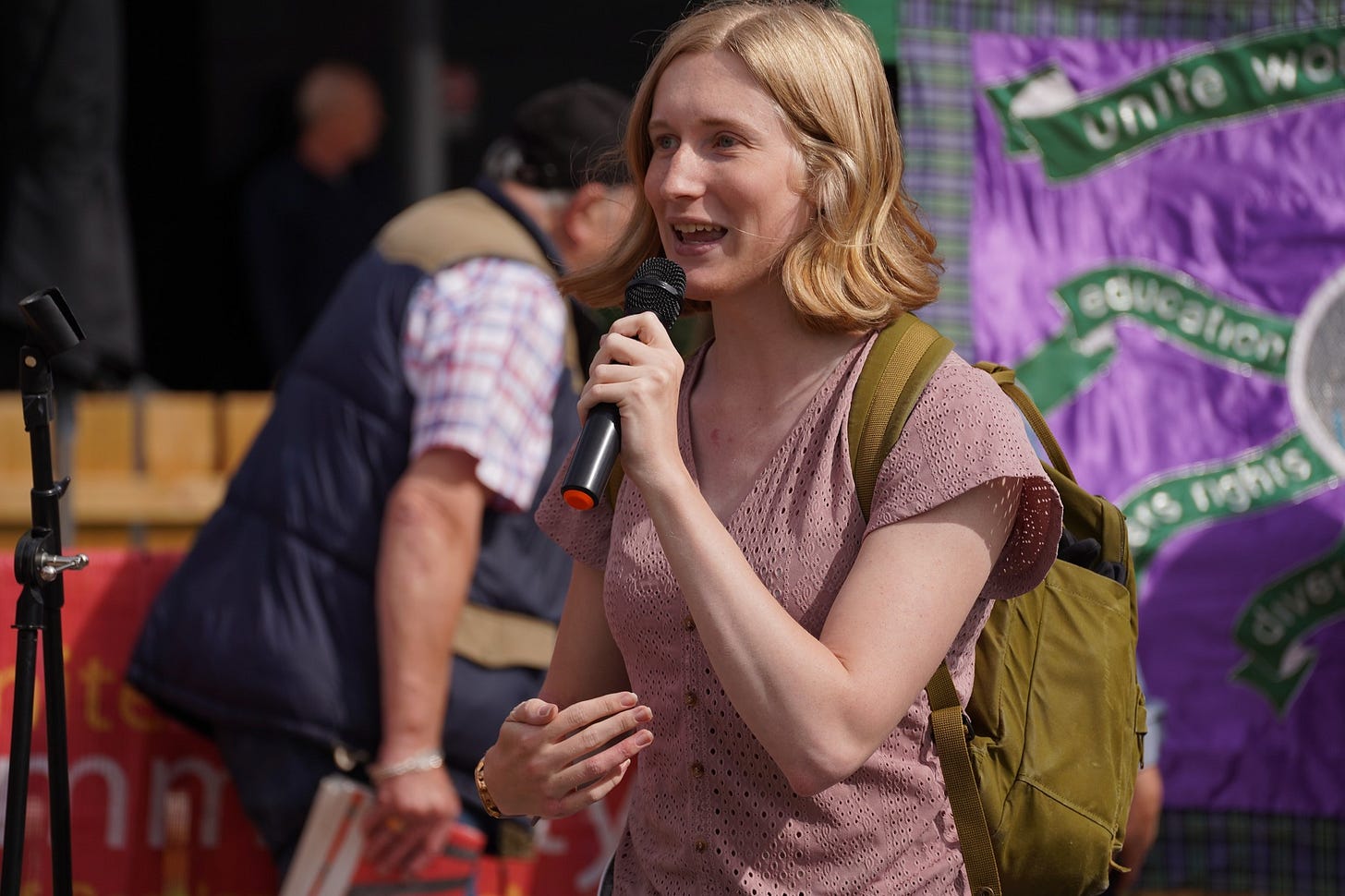 Ellie speaking at the Unite picket. She wears a pink blouse, has a green backpack and holds a microphone.