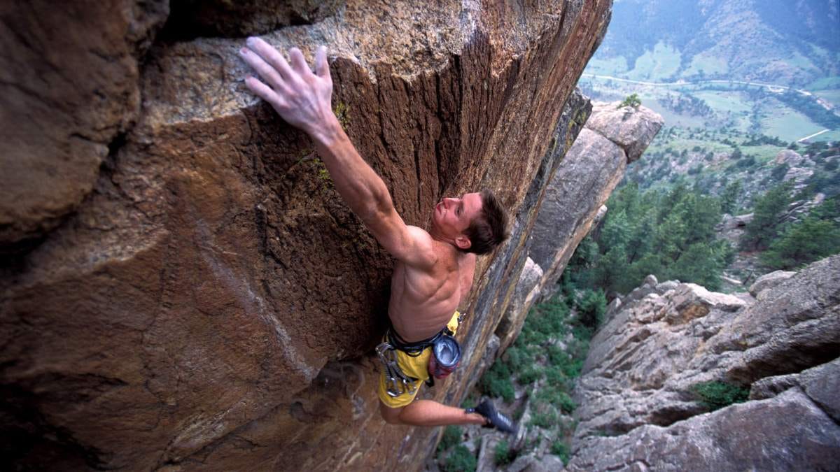 A shirtless man, resembling Tommy Caldwell, climbs a steep rock face high above the ground, reaching for a hold with one hand and gripping the rock with the other; trees and a valley stretch out below. A shirtless man, resembling Tommy Caldwell, climbs a steep rock face high above the ground, reaching for a hold with one hand and gripping the rock with the other; trees and a valley stretch out below.