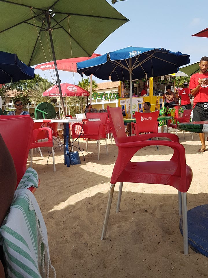 A mojito with mint and lime on a beachside table, next to a small plastic beach bar with outdoor seating set directly on the sand.