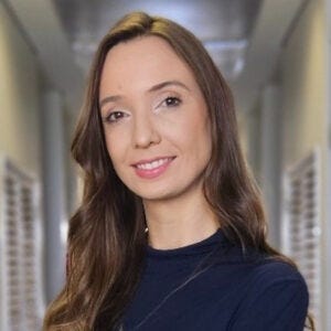 A imagem mostra um retrato em plano médio (da cintura para cima) de uma mulher jovem, com pele clara e cabelos castanhos claros, longos e ondulados, que caem sobre seu ombro direito. Ela está posicionada no centro da foto, levemente voltada para a esquerda, mas com o rosto e o olhar direcionados para a câmera, exibindo um sorriso suave e gentil. Ela veste uma blusa de gola alta e mangas compridas na cor azul-marinho. Seus braços estão cruzados à frente do corpo.