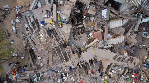 Aerial drone view of a multi-story building reduced to rubble after the earthquake, with rescue workers in helmets and hi-vis gear searching through the collapsed floors and debris. A concrete staircase remains partially intact on the right; scattered buckets, twisted rebar, and wreckage cover the site. Photo: Reuters.