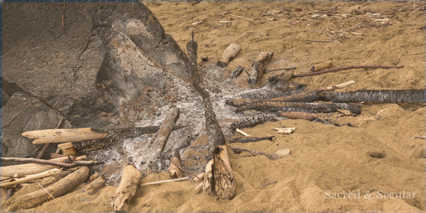 The remains of a charcoal campfire on a sandy beach beside large rocks, with driftwood scattered around the ash. The fire has burned low, creating a quiet, reflective scene that evokes memory, restoration and the hope of a new beginning.