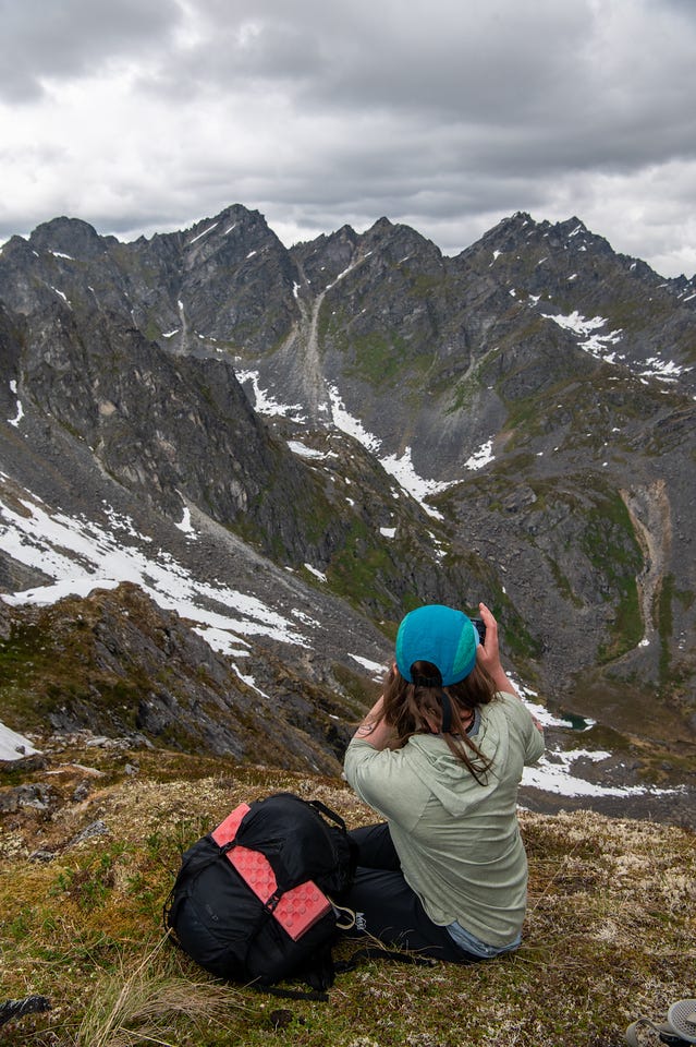 A hiker sits on the tundra at the edge of a pass, seen from behind in a pale green hoody and teal cap, holding up a phone to photograph the view. Her black pack with pink accents lies beside her. Below and beyond, a wall of jagged grey peaks rises above a valley holding long snowfields, a small tarn just visible far below, under a heavy overcast sky. A hiker sits on the tundra at the edge of a pass, seen from behind in a pale green hoody and teal cap, holding up a phone to photograph the view. Her black pack with pink accents lies beside her. Below and beyond, a wall of jagged grey peaks rises above a valley holding long snowfields, a small tarn just visible far below, under a heavy overcast sky.