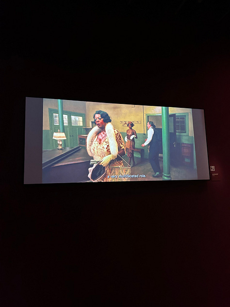 Viola Davis on a screen, costumes on mannequins, Marlee Matlin accepting her Oscar, an Oscar in a case, and photos of the Black Panthers from a documentary.