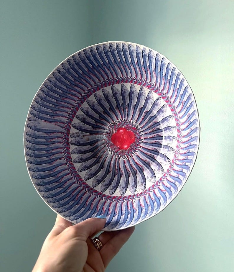 May include: A round ceramic bowl featuring a repeating pattern of blue and silver fish arranged in concentric circles. The center of the bowl displays a bright red, glossy, raised circular accent piece that contrasts with the fish motif.