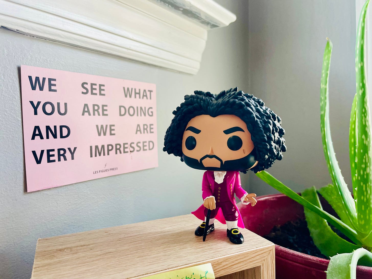 [alternate description: Funko Pop of Daveed Digs in Hamilton rests on a small wooden table next to a light pink sign that reads “We See What You Are Doing and We Are Very Impressed.” An aloe plant bends into the right side of the photo