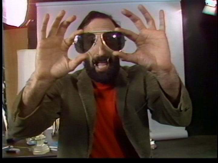 From The Pressures of the Text: Peter Rose stands on a set furnished like a professor's office, addresses the viewer and holds a pair of sunglasses up in front of his head so it almost looks like he's wearing them