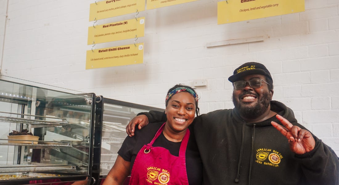 Two people - Mahalia Chambers and Jowayne Marks - standing in front of a metal canteen with a yellow sign hanging above them