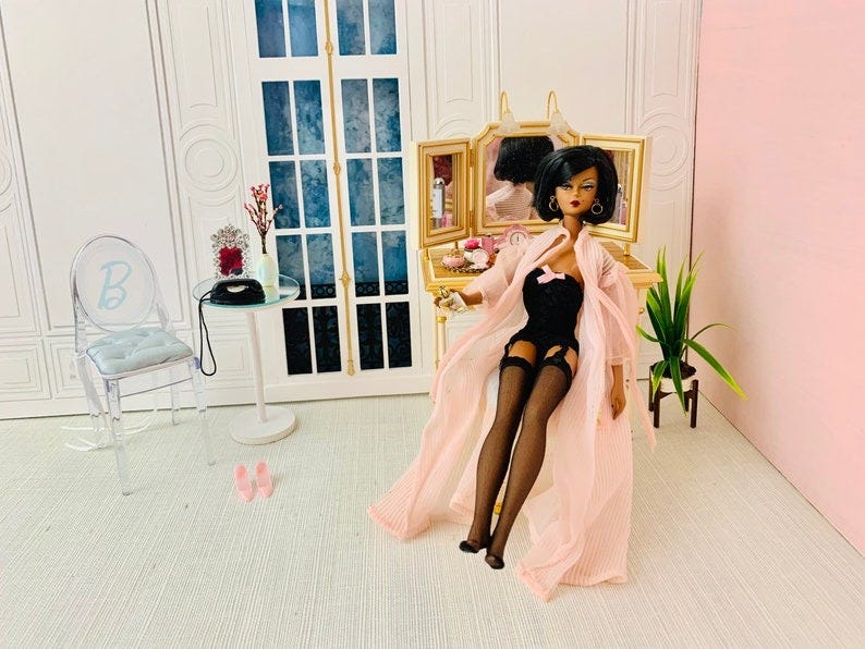 May include: A doll wearing a pink robe and black lingerie sitting at a vanity with a triple mirror. The vanity has a gold frame and is decorated with a small bouquet of red flowers and a clock. The doll has black hair and is wearing black stockings and heels. The doll is sitting on a white chair with a blue letter B on the back. The doll is looking at the mirror. There is a small table with a black purse and a plant in the background.