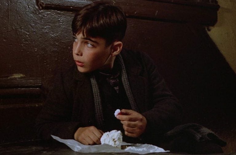 Frame da "C'era una volta in America" con Patsy, che è un ragazzino di 8-9 anni dalla pelle chiara, seduto su delle scale interne a un palazzo con le pareti marroni. Sta guardando verso destra qualcosa fuori dall'inquadratura, mentre regge nella mano sinistra una charlotte di panna.