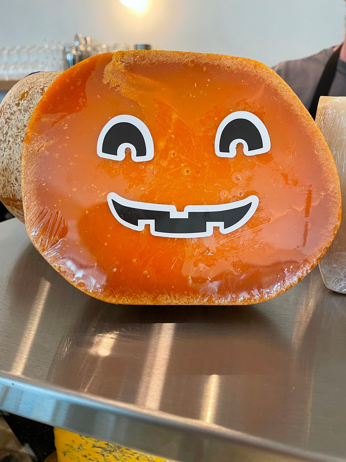 A half wheel of Mimolette with stickers of eyes and a mouth, making it look like a Jack-o‘Lantern A half wheel of Mimolette with stickers of eyes and a mouth, making it look like a Jack-o‘Lantern