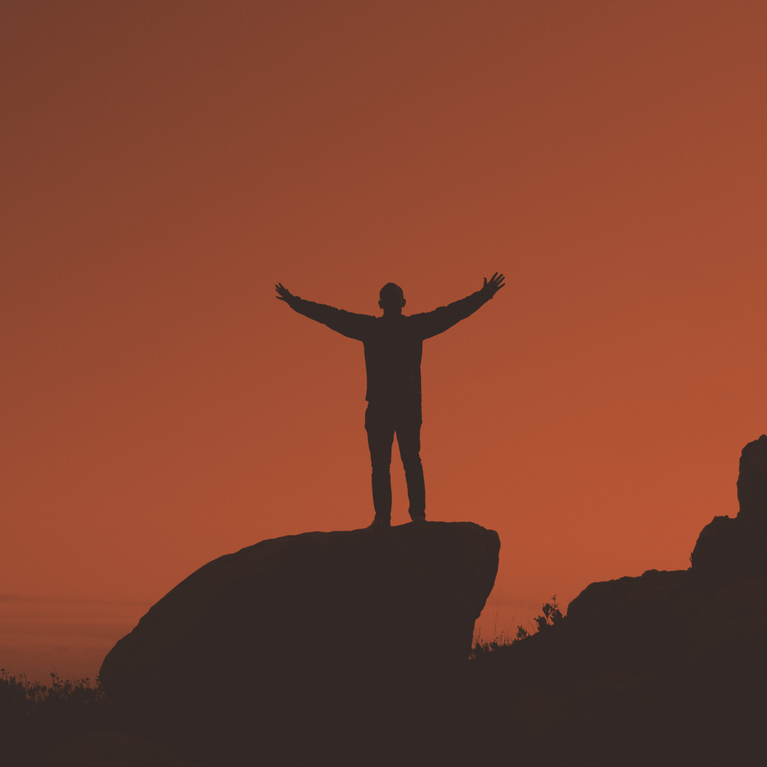 A person stands triumphantly with their arms spread wide atop a rock.