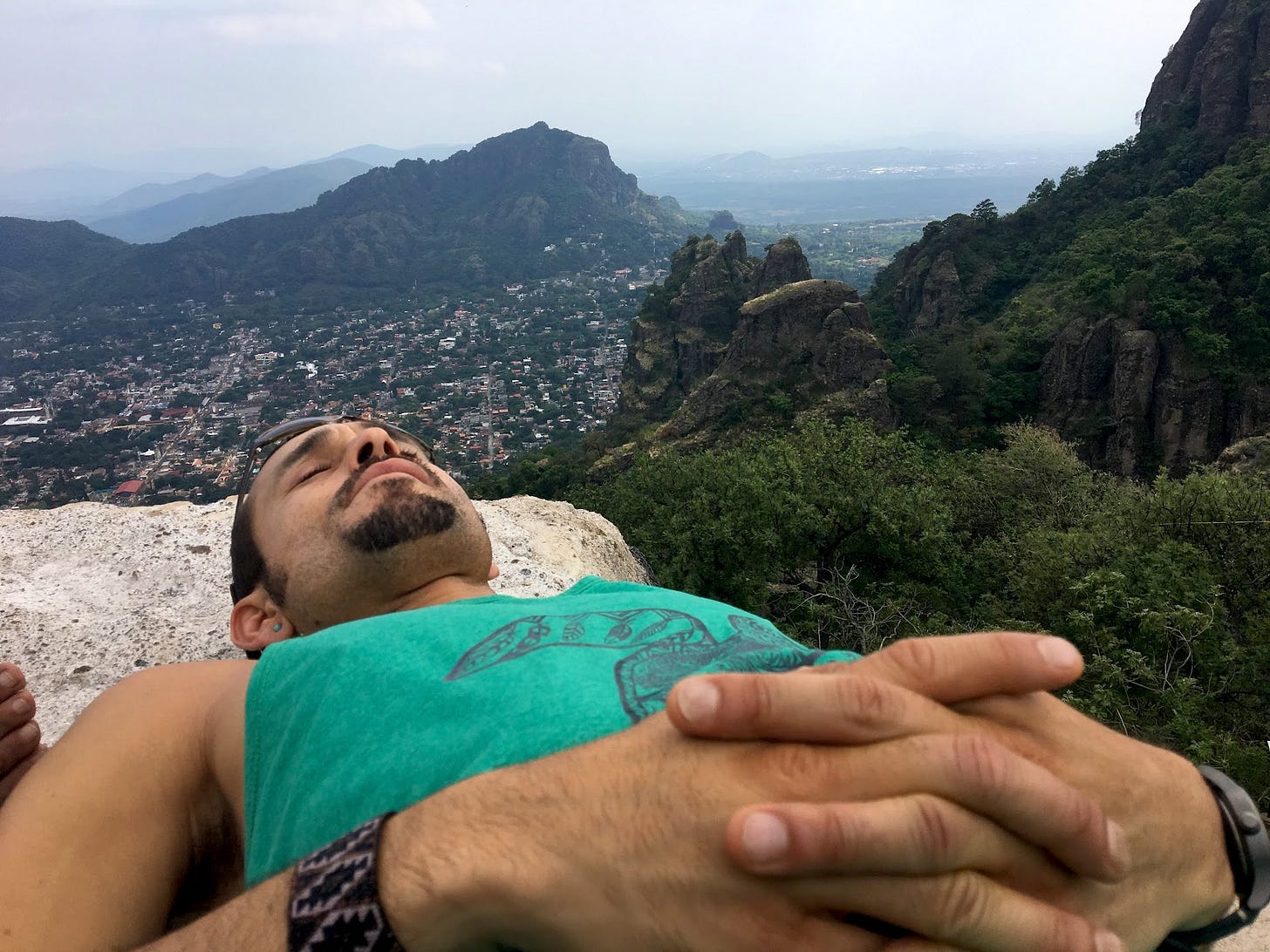 Gera is lying on a large flat rock atop a mountain, with his eyes closed in meditation. Other mountains are visible in the distance.