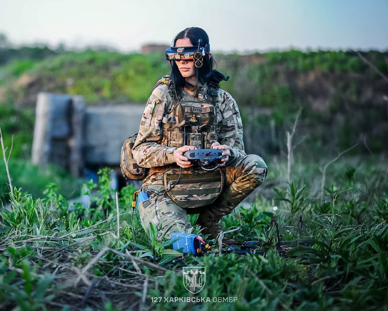 A Ukrainian soldier in camouflage kneels in a field wearing first-person-view goggles and holding a drone controller, with an assembled quadcopter on the ground in front of her. A Ukrainian soldier in camouflage kneels in a field wearing first-person-view goggles and holding a drone controller, with an assembled quadcopter on the ground in front of her.