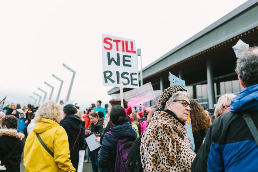photo of a woman's march. photo is centered on a sign that reads "Still We Rise"