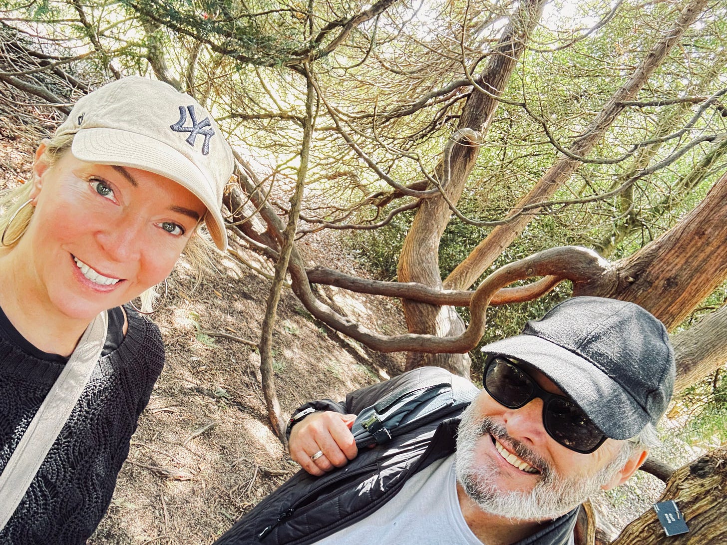Kelly and Nigel smiling under a canopy of twisting tree branches on a sunny day during a slow-travel walk.