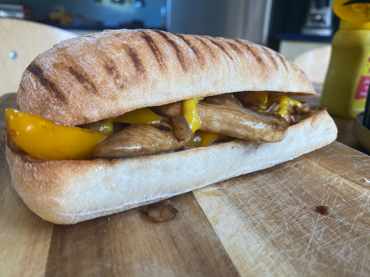 Wooden board with a sandwich on top. The filling is mushrooms and peppers. A bottle of American mustard is in the background.