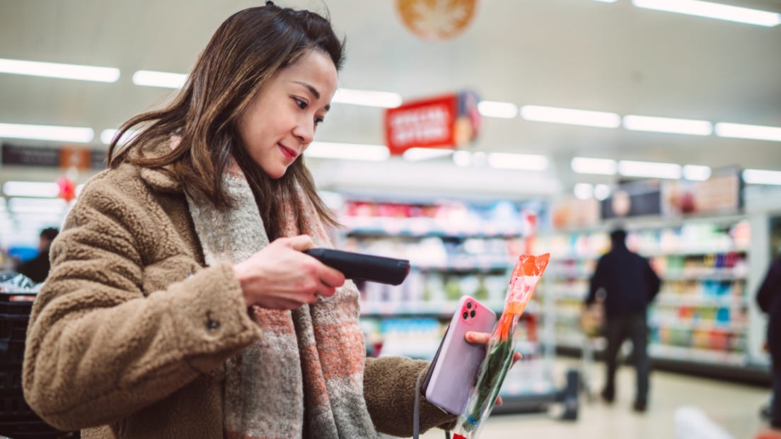 Young woman with dark hair, wearing brown fake fur jacket and grey and pink scarf scans packet of herbs at supermarket Young woman with dark hair, wearing brown fake fur jacket and grey and pink scarf scans packet of herbs at supermarket