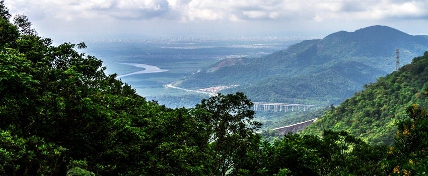 Vista da Baixada Santista do alto da Serra do Mar. Em primeiro plano, morros cobertos por vegetação de mata atlântica. No centro da foto, ao longe, um aglomerado de cidades, e estradas e cursos d'água preenchendo o caminho entre a serra e a cidade