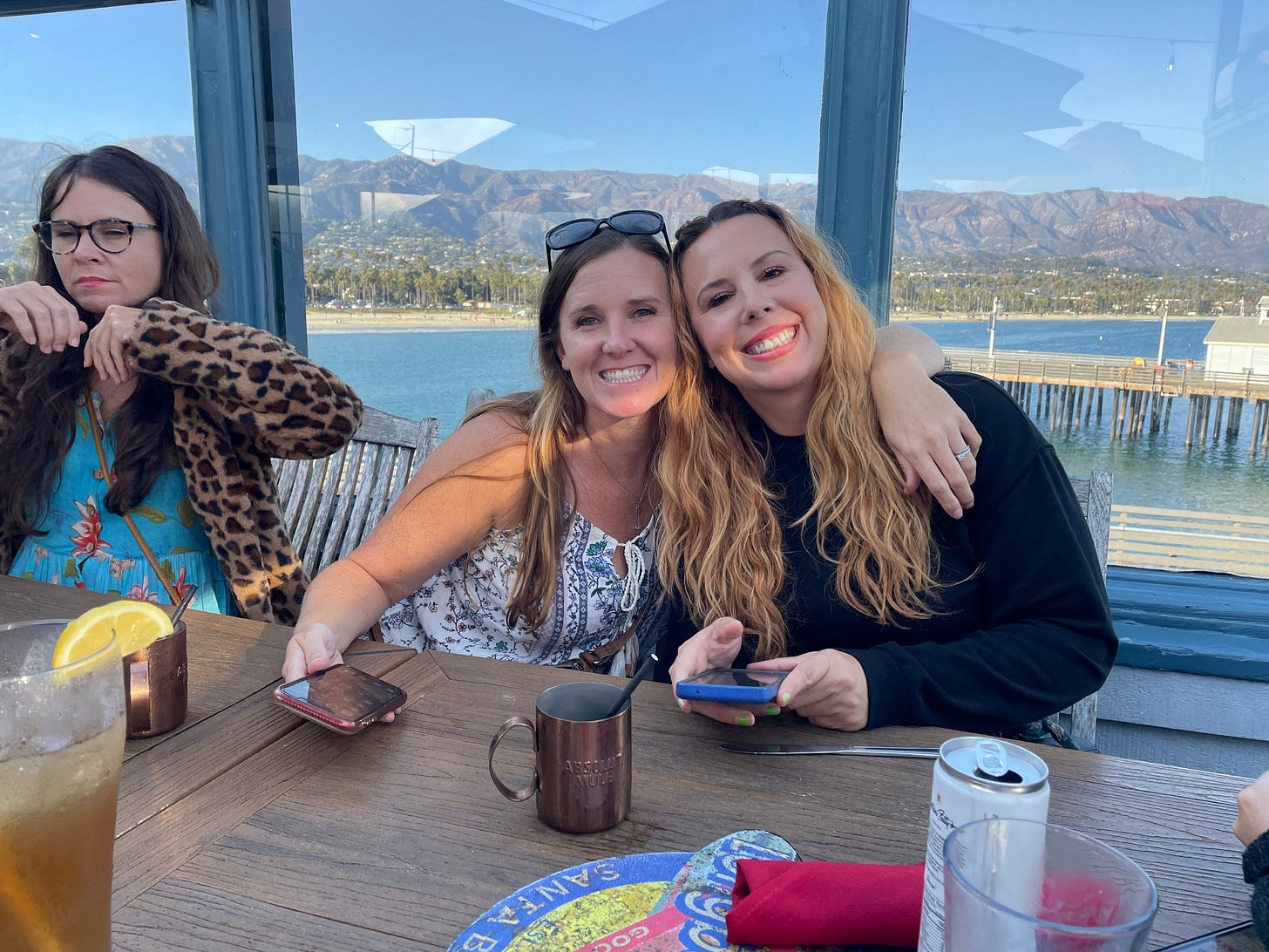Three women at a table. One has eyes half closed and looks sleepy and dorky. The other two have pretty smiles. Background view of a sunny beach.