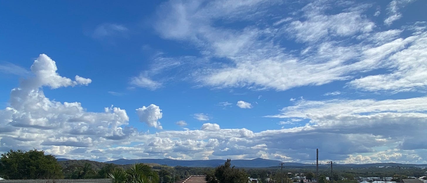 Blue sky and clouds with distant views of a mountain, trees and rooftops in the forground with telegraph poles now leafless dreaming of a time the were free to grow and not work all day.